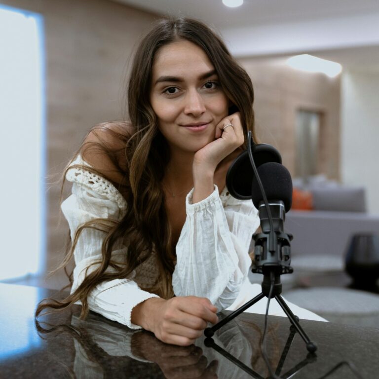 A woman with long brown hair rests her chin in her hand and smiles behind a podcast microphone.