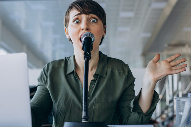 A woman with short brown hair sits behind a desk microphone, her eyebrows raised and eyes wide in an expression of overwhelm.