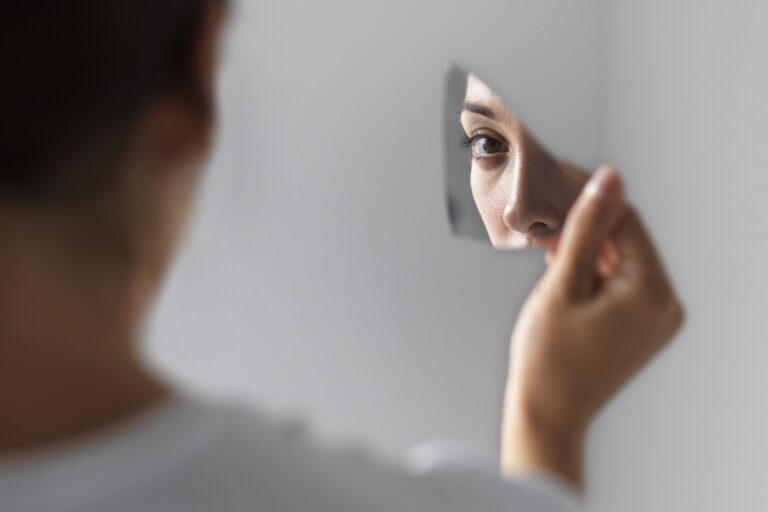 A woman holds a piece of broken mirror, looking at her reflection and contemplating her imposter syndrome around podcasting.