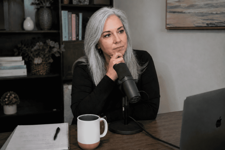A middle-aged woman with long grey hair sits at a podcast microphone, chin resting on hand, looking thoughtful and hesitant.