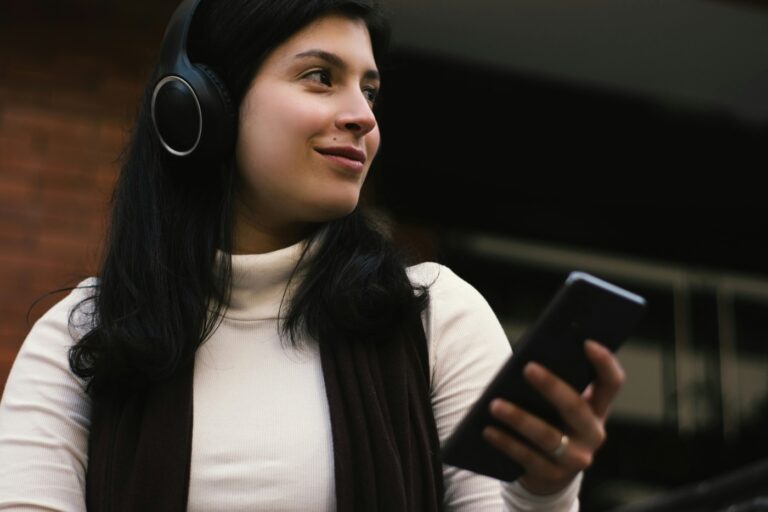 A woman with long, dark hair wearing a white turtleneck and headphones listens to an audio-first podcast on her smartphone.